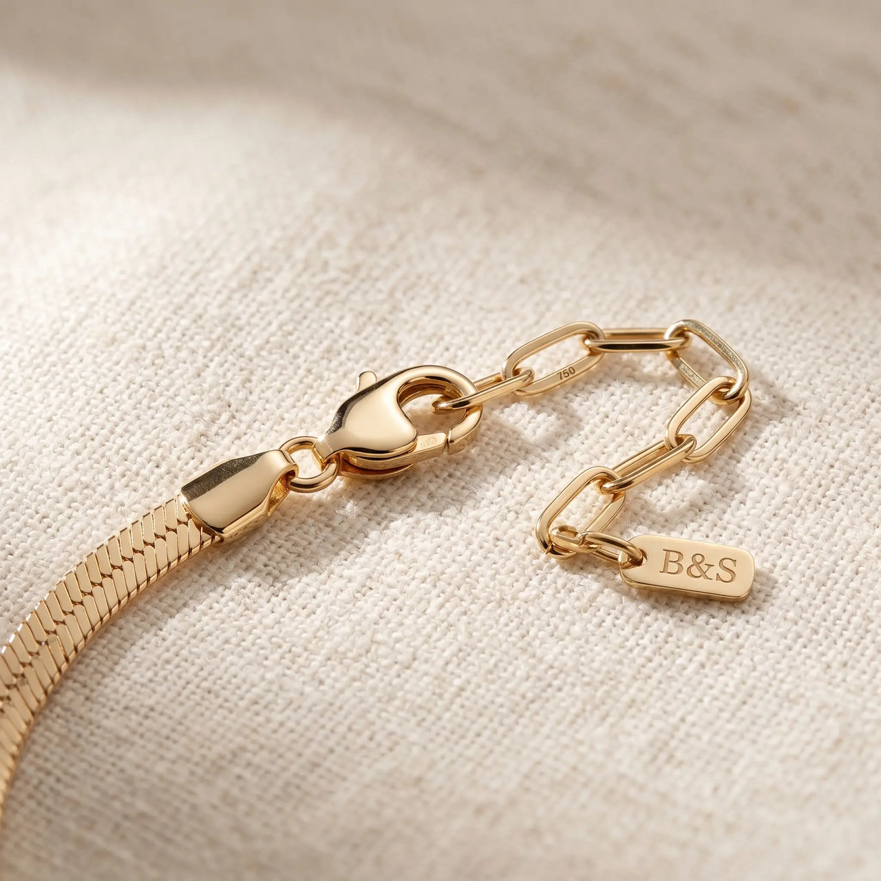 Layla gold anklet macro detail — lobster clasp + paperclip extension chain + B&S engraved metal tag on cream linen